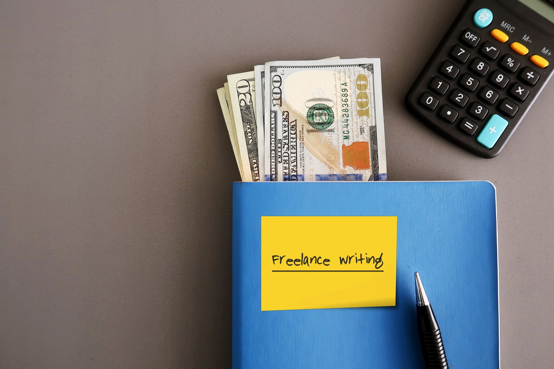 Cash dollars money, calculator , a pen, yellow note on a blue notebook written FREELANCE WRITING , on grey background, concept of financial planning, make extra income from freelance writing side job